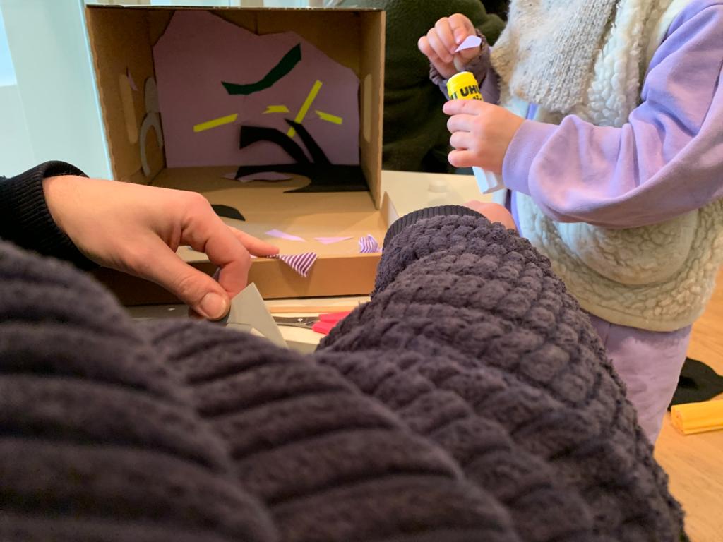 Ein Kind und eine erwachsene Person bekleben den Innenteil einer Pappkiste mit Papier und Stoff.