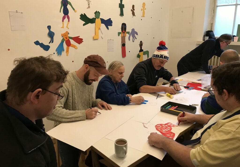Workshopteilnehmende sitzen an einem langen Tisch und arbeiten künstlerisch. An der Wand hängen kleine Figuren aus Papier, die von den Teilnehmenden gestaltet wurden.