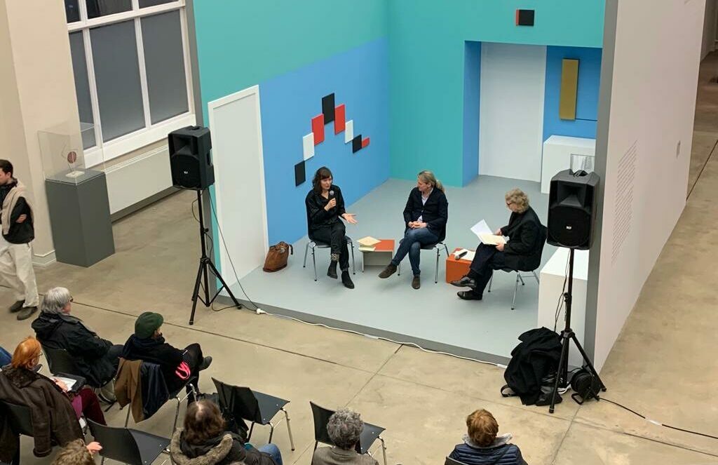 Ansicht auf eine Veranstaltung in der Ausstellung von Erich Buchholz. Auf einem Podium sitzen drei Personen, die sich unterhalten. Ihnen gegenüber sitzt das Publikum auf Stühlen.